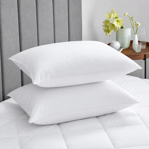 Two white pillows on a bed with a gray headboard and a small table with flowers in the background.