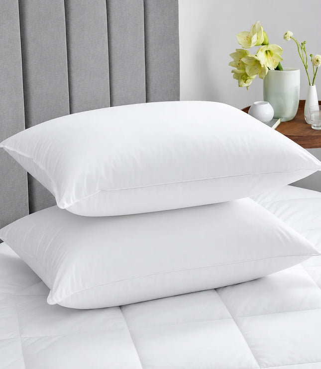 Two white pillows on a bed with a gray headboard and a small table with flowers in the background.